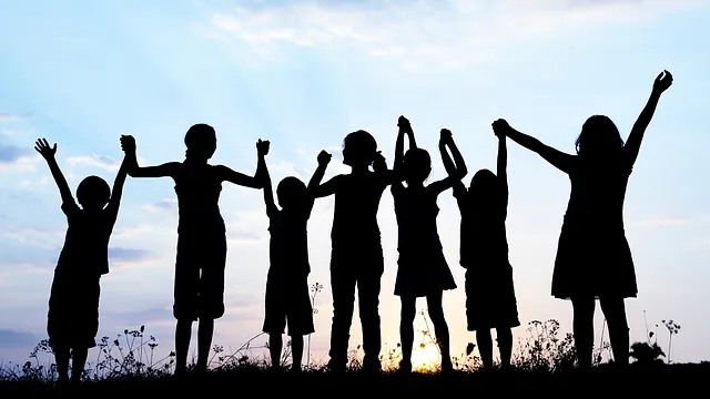 Silhouetten von Kindern, die Hände fassend in einer Reihe stehen und ihre Hände in die Luft jubelnd werfen vor einem Sonnenuntergang
