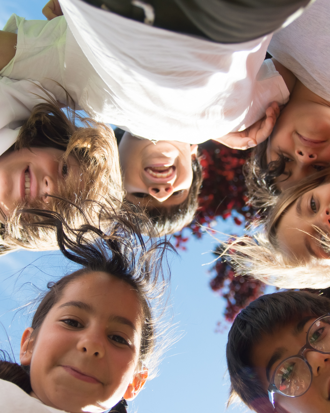 Kinder stehen im umarmend im Kreis und richten ihre Köpfe zum Boden, wo die Kamere in der Mitte vom Kreis liegt